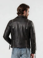 Ein Mann mit braunem Haar steht mit dem Rücken zur Kamera und trägt die trueprodigy Biker Lederjacke Herren "Jackson" mit Schnallen und hellblaue Jeans vor einem schlichten weißen Hintergrund.