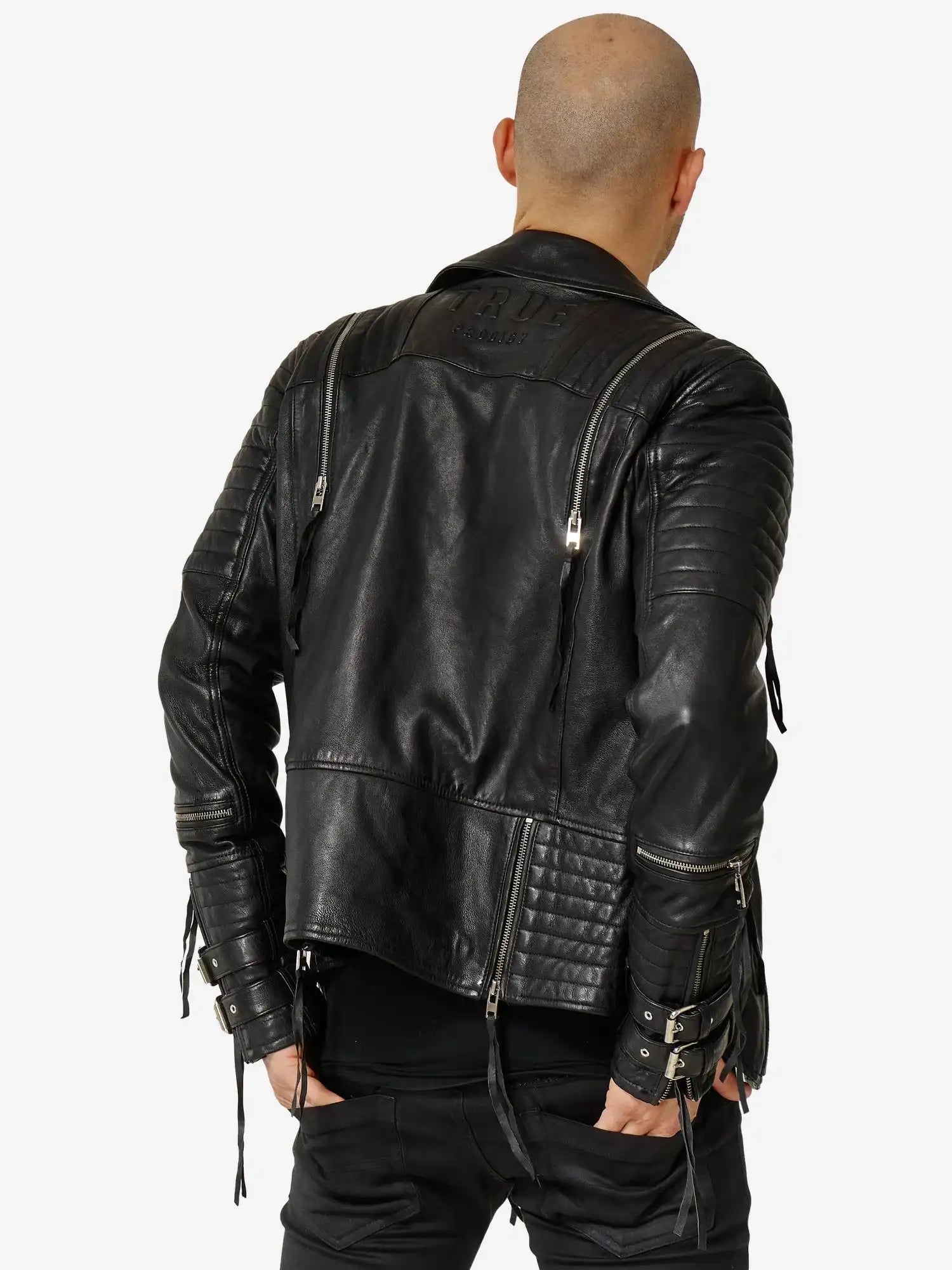 Eine Person mit rasiertem Kopf wird von hinten in einer schwarzen Jackson Bikerlederjacke mit Schnallen von trueprodigy, gepaart mit einer schwarzen Hose, vor einem schlichten weißen Hintergrund gezeigt.
