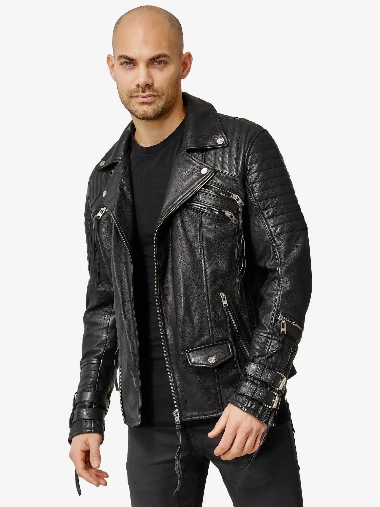 Ein Mann mit rasiertem Kopf und gestutztem Bart trägt eine schwarze Trueprodigy Jackson Bikerlederjacke mit Schnallen über einem schwarzen Hemd, das mit silbernen Reißverschlüssen und Schnallen versehen ist, vor einem schlichten weißen Hintergrund.