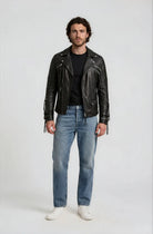 Ein Mann mit mittellangem Haar und Bart steht mit dem Gesicht nach vorne in der trueprodigy Biker Lederjacke Herren "Jackson" mit Schnallen, gepaart mit einem schwarzen T-Shirt, hellblauen Jeans und weißen Turnschuhen vor einem schlichten hellen Hintergrund.