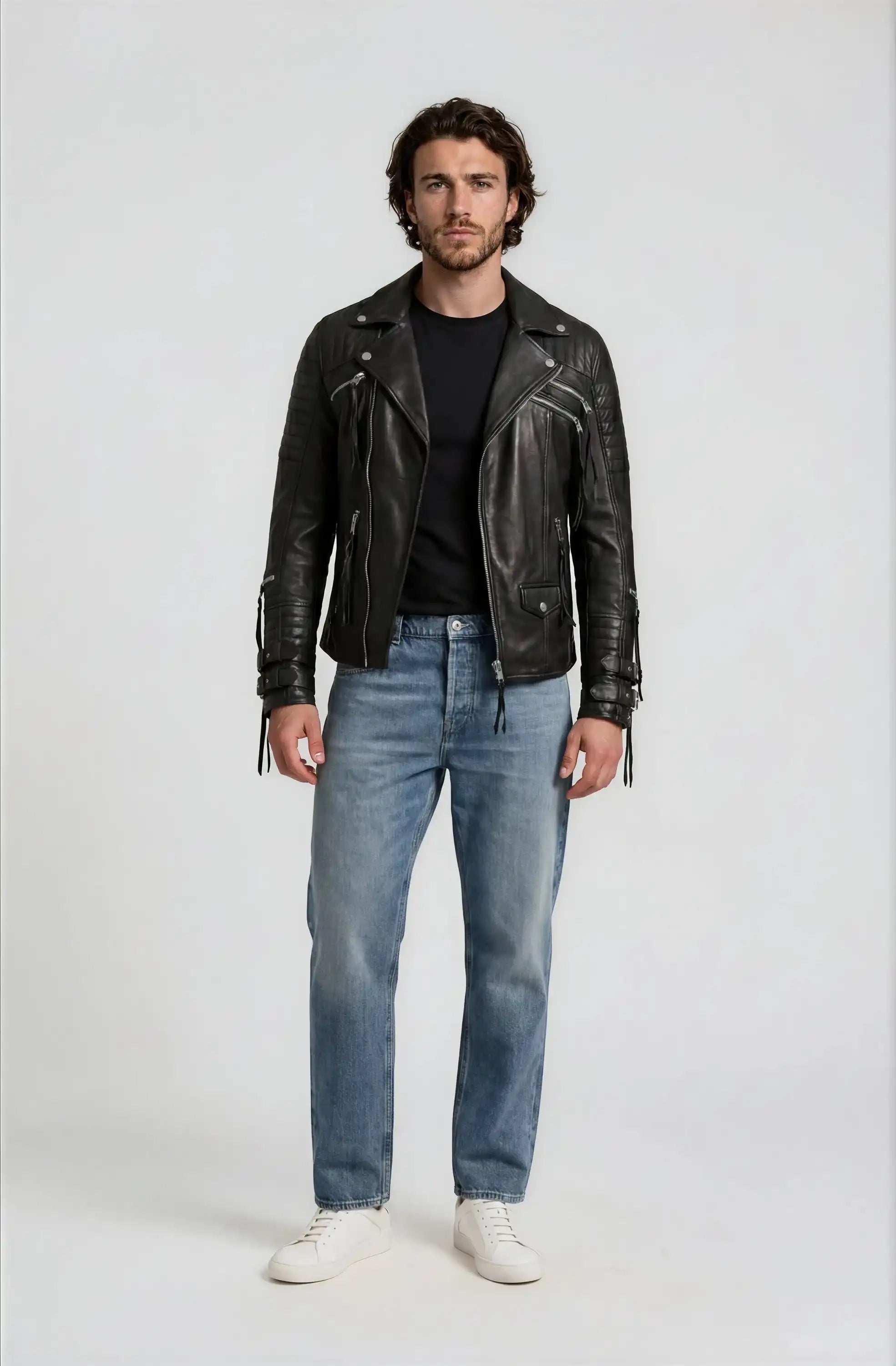 Ein Mann mit mittellangem Haar und Bart steht mit dem Gesicht nach vorne in der trueprodigy Biker Lederjacke Herren "Jackson" mit Schnallen, gepaart mit einem schwarzen T-Shirt, hellblauen Jeans und weißen Turnschuhen vor einem schlichten hellen Hintergrund.