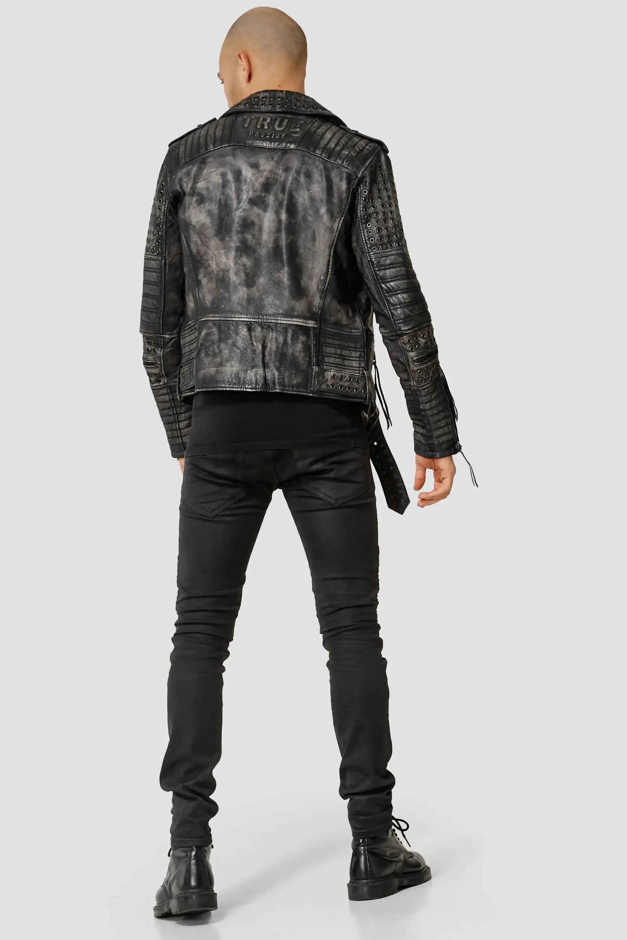 Eine Person mit rasiertem Kopf wendet sich von der Kamera ab und trägt die Geralt Vintage Lederjacke mit Ösen von trueprodigy, gepaart mit schwarzen Skinny-Hosen und Stiefeln, vor einem schlichten hellgrauen Hintergrund.