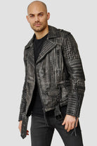 Ein Mann mit rasiertem Kopf und kurzem Bart trägt eine echte Geralt Vintage Lederjacke mit Ösen - eine kantige schwarze Lederjacke - über einem schwarzen Hemd und einer Hose vor einem schlichten hellgrauen Hintergrund.