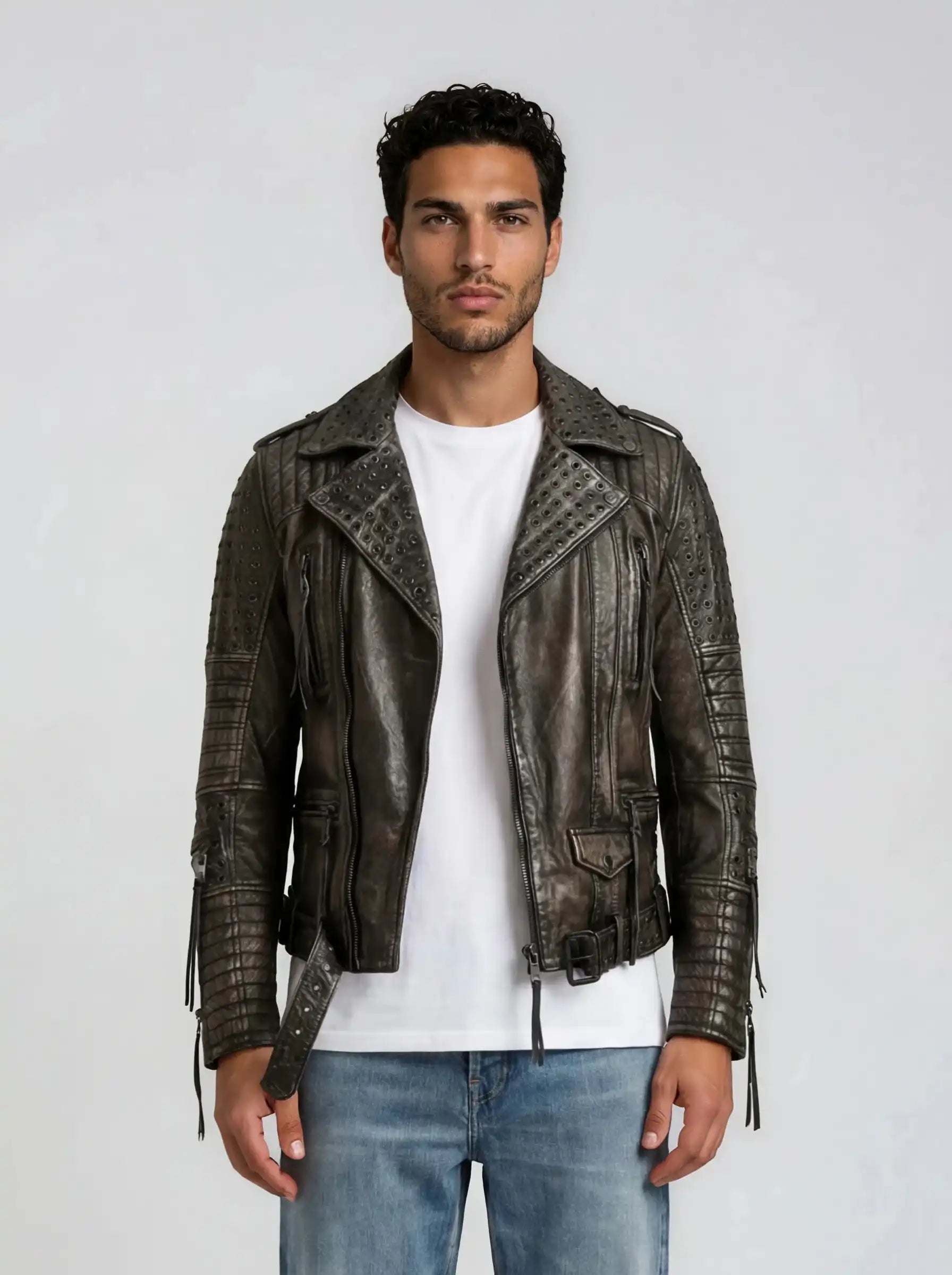 Ein Mann mit kurzen lockigen Haaren trägt ein weißes T-Shirt, blaue Jeans und die trueprodigy Vintage Biker Lederjacke Herren "Geralt" mit Ösen. Er steht mit dem Gesicht nach vorne vor einem schlichten hellgrauen Hintergrund.