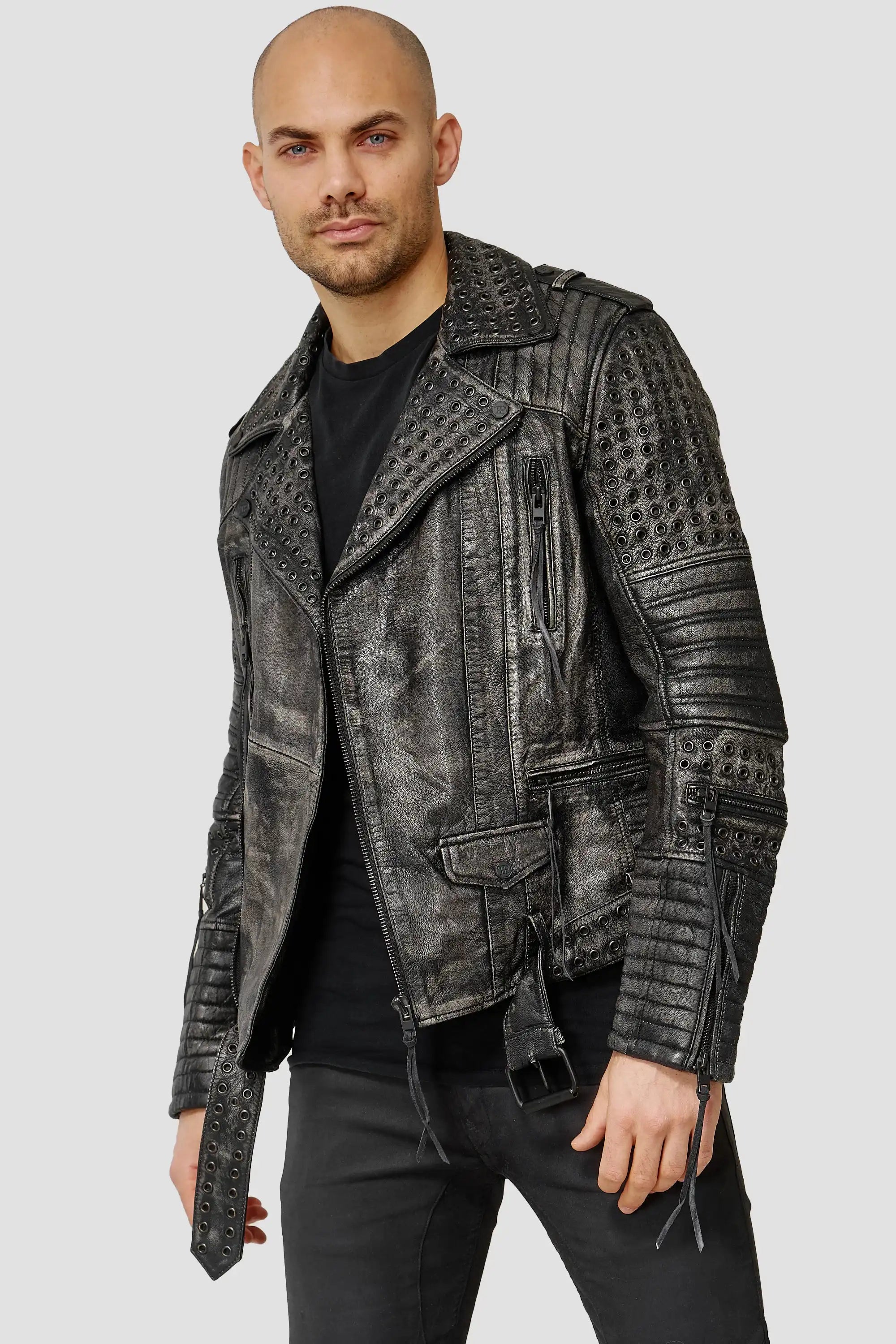 Ein Mann mit rasiertem Kopf und kurzem Bart trägt eine echte Geralt Vintage Lederjacke mit Ösen - eine kantige schwarze Lederjacke - über einem schwarzen Hemd und einer Hose vor einem schlichten hellgrauen Hintergrund.
