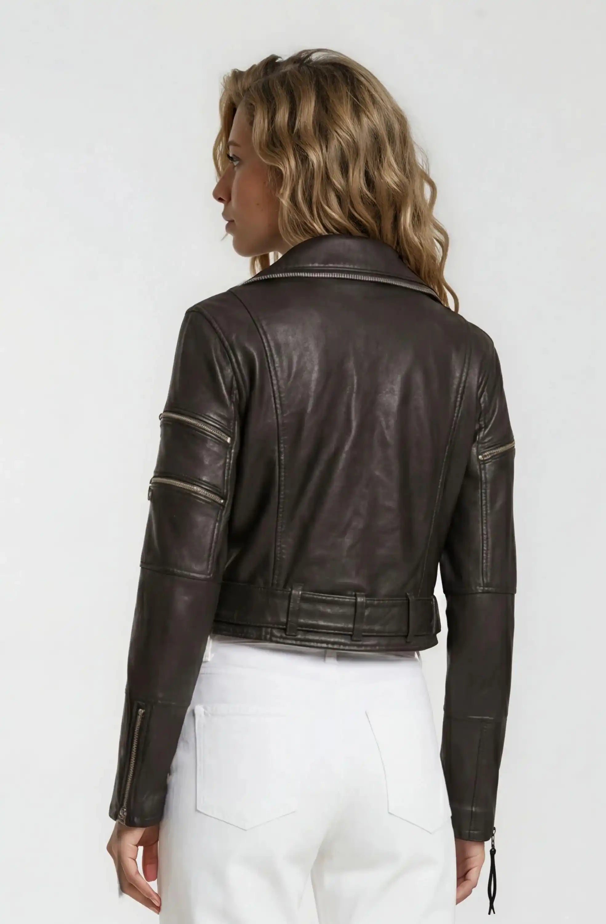 Eine Person mit gewelltem blondem Haar ist von hinten zu sehen. Sie trägt eine Trueprodigy Bikerlederjacke mit Gürtel mit Reißverschluss und eine weiße Hose vor einem schlichten hellen Hintergrund.