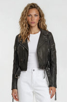 Eine Frau mit welligem blondem Haar trägt die Bikerlederjacke mit Gürtel von trueprodigy über einem weißen T-Shirt und hoch taillierten weißen Hosen vor schlicht hellem Hintergrund.