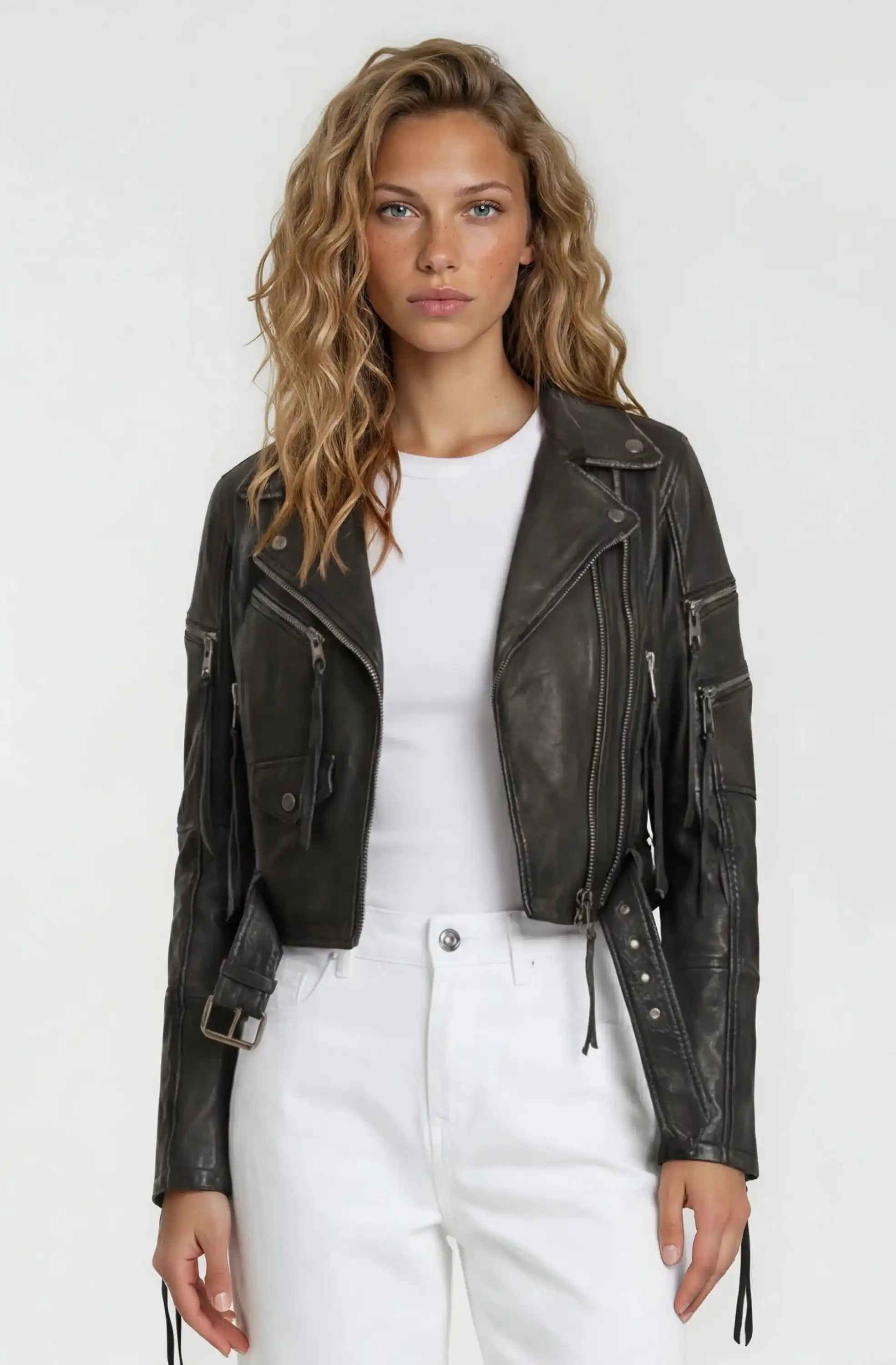 Eine Frau mit welligem blondem Haar trägt die Bikerlederjacke mit Gürtel von trueprodigy über einem weißen T-Shirt und hoch taillierten weißen Hosen vor schlicht hellem Hintergrund.