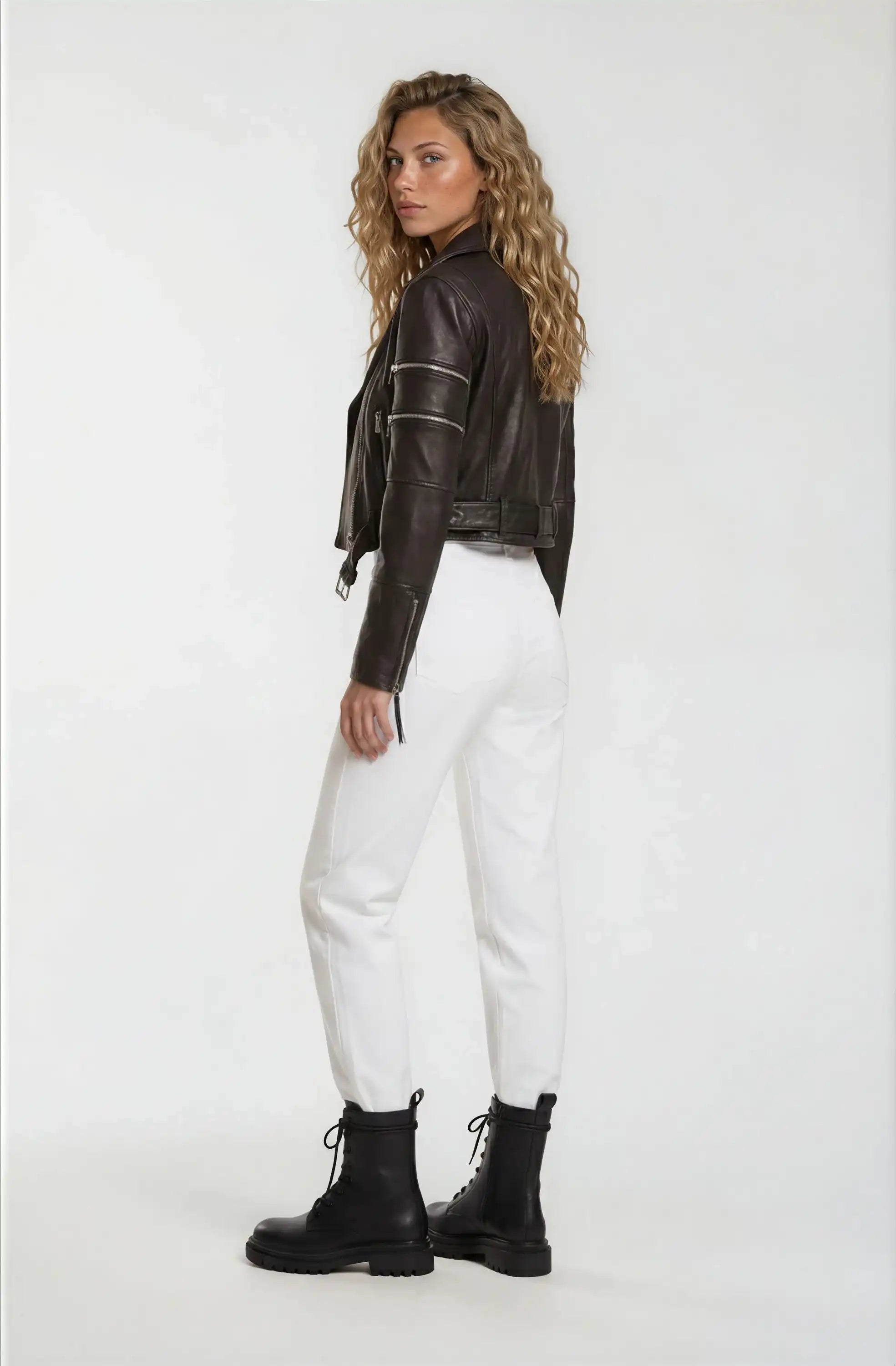 Eine Frau mit gewelltem blondem Haar steht leicht gedreht und trägt die trueprodigy Bikerlederjacke mit Gürtel aus Schafsleder, weißen Hosen und schwarzen Schnürstiefel vor einem schlichten weißen Hintergrund.