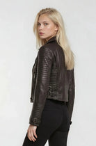 Eine Frau mit langen blonden Haaren steht leicht zur Seite gedreht, trägt eine trueprodigy Bikerlederjacke mit Schnallen und eine schwarze Hose und blickt über ihre Schulter in die Kamera vor einem schlichten hellen Hintergrund.