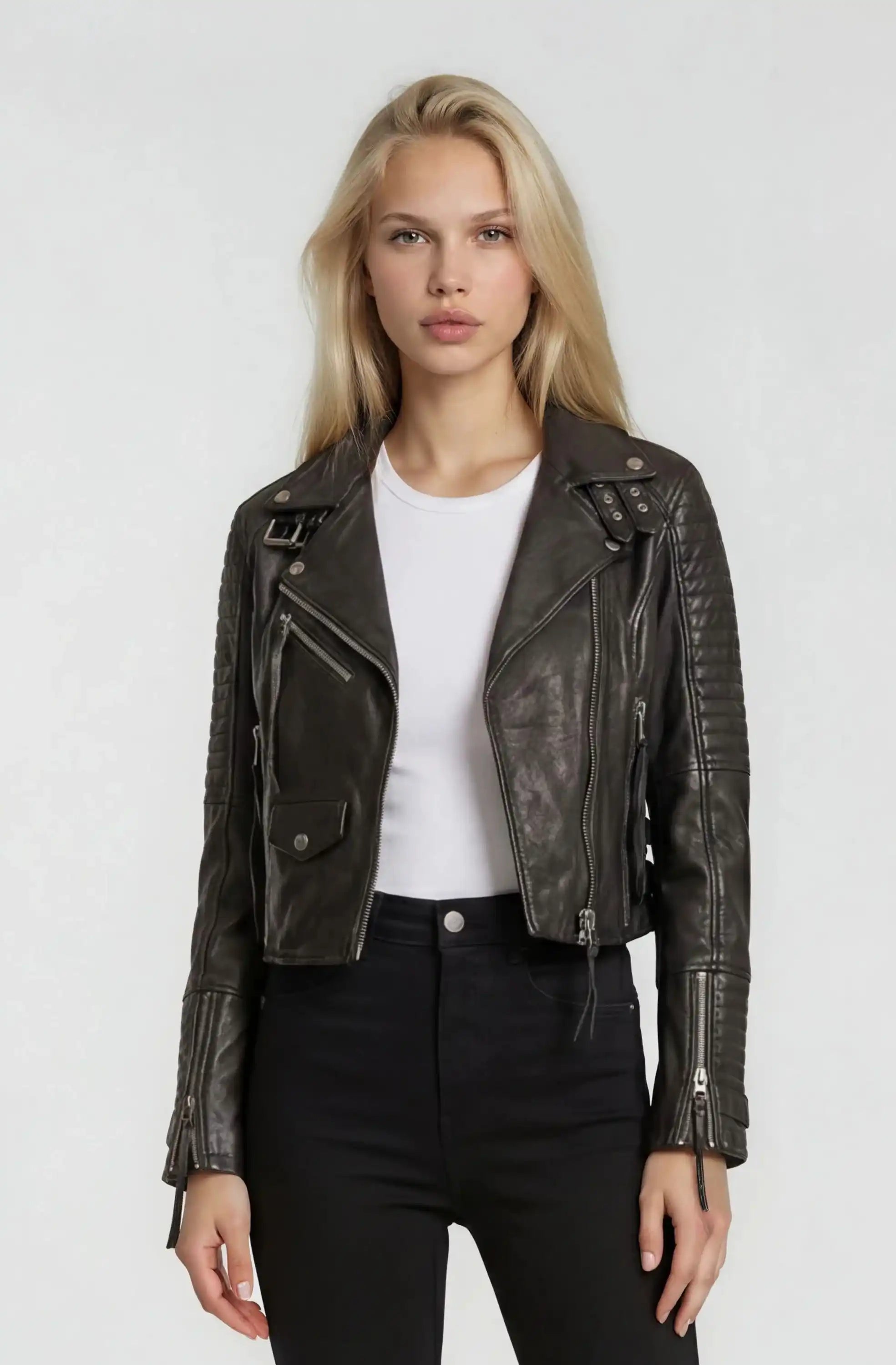 Eine Person mit langen blonden Haaren trägt die trueprodigy Bikerlederjacke mit Schnallen im klassischen Biker-Look über einem weißen T-Shirt und einer hochgezogenen schwarzen Hose, stehend vor einem schlichten hellen Hintergrund.