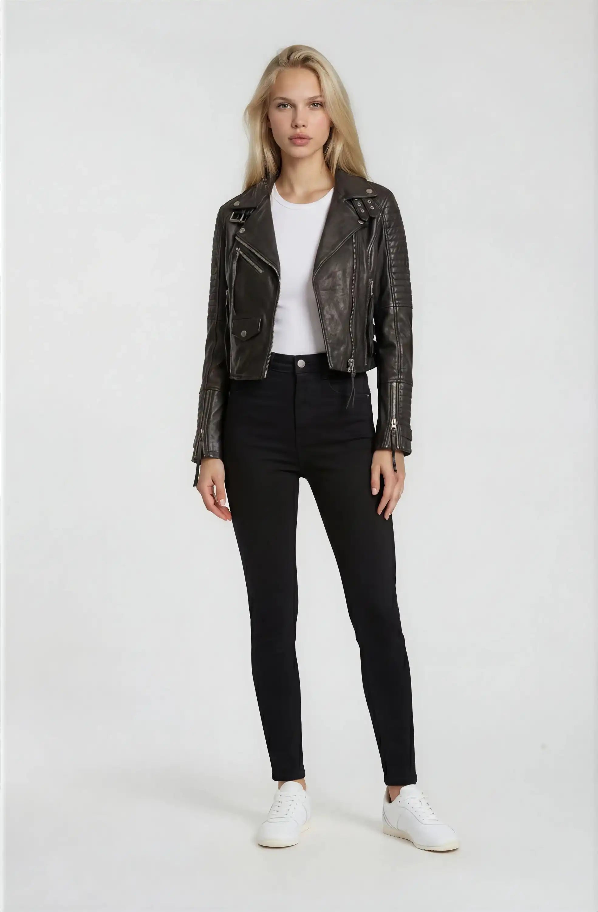 Eine Person mit langen blonden Haaren steht vor einem schlichten Hintergrund und trägt die Bikerlederjacke mit Schnallen von trueprodigy, gepaart mit einem weißen T-Shirt, einer schwarzen High-Waist-Hose und weißen Sneakers.