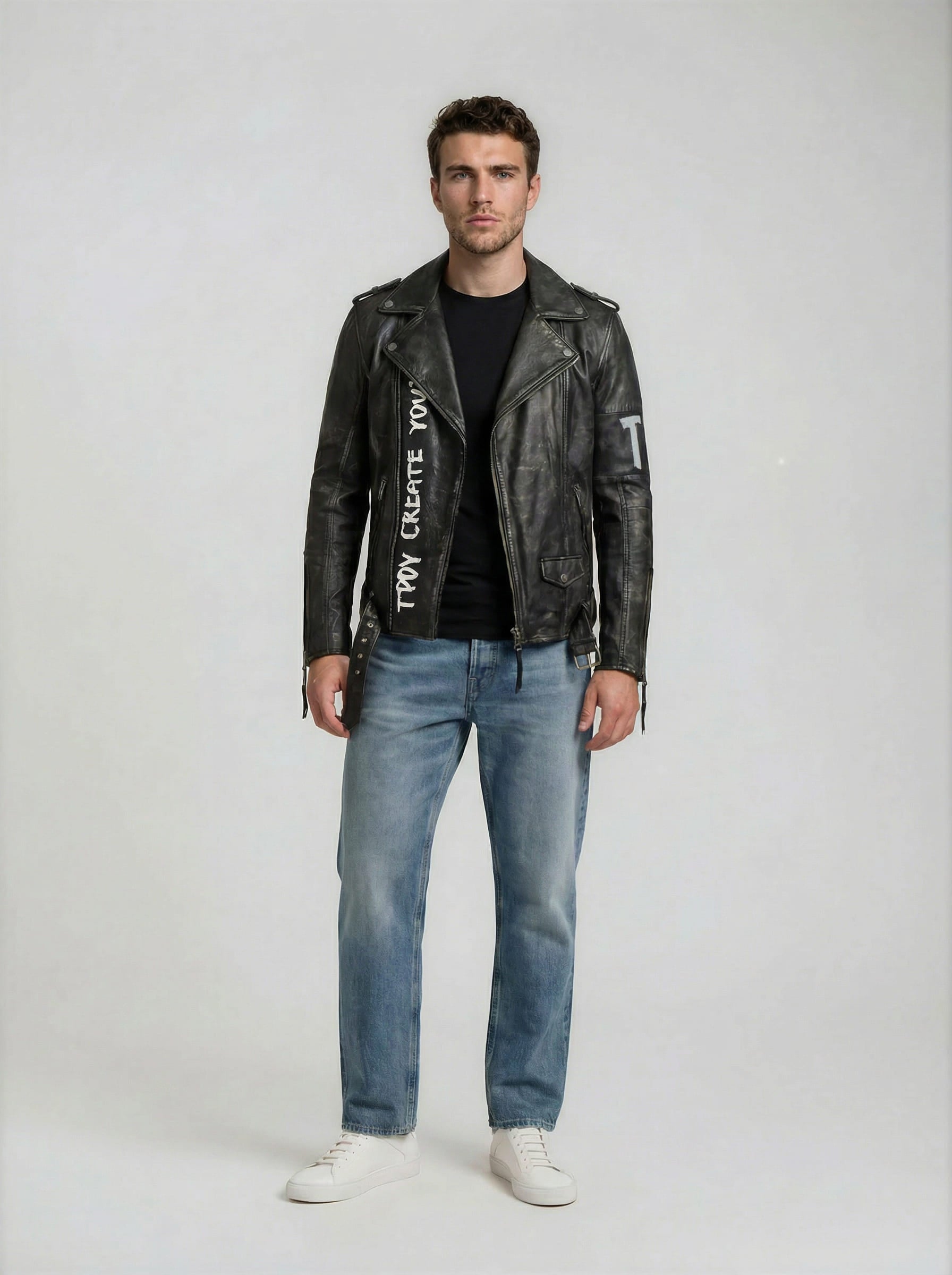 In der trueprodigy Biker Lederjacke Herren "Russell" mit Rücken-Print, gepaart mit einem schwarzen T-Shirt, hellblauen Jeans und weißen Turnschuhen, blickt ein Mann nach vorne und sorgt vor einem schlichten Hintergrund für einen coolen Biker-Look.