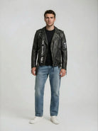 In der trueprodigy Biker Lederjacke Herren "Russell" mit Rücken-Print, gepaart mit einem schwarzen T-Shirt, hellblauen Jeans und weißen Turnschuhen, blickt ein Mann nach vorne und sorgt vor einem schlichten Hintergrund für einen coolen Biker-Look.