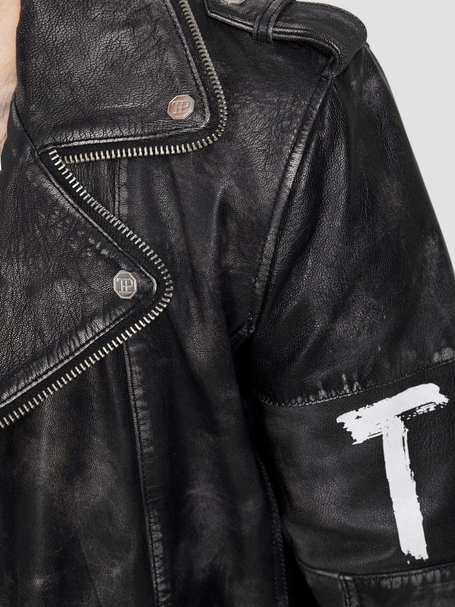 Nahaufnahme der trueprodigy Russell Lederjacke mit Print: schwarzes Ziegenleder, Metalldruckknöpfe, freiliegender silberner Reißverschluss, weiß lackiertes T auf dem Ärmel, und ein strukturierter, leicht abgenutzter Biker-Look.