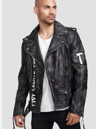 Ein Mann in der trueprodigy Russell Lederjacke mit Print - einer schwarzen Lederjacke im Biker-Stil mit weißem Graffiti-Schriftzug -, einem weißen T-Shirt und einer schwarzen Hose steht vor einem schlichten hellgrauen Hintergrund.
