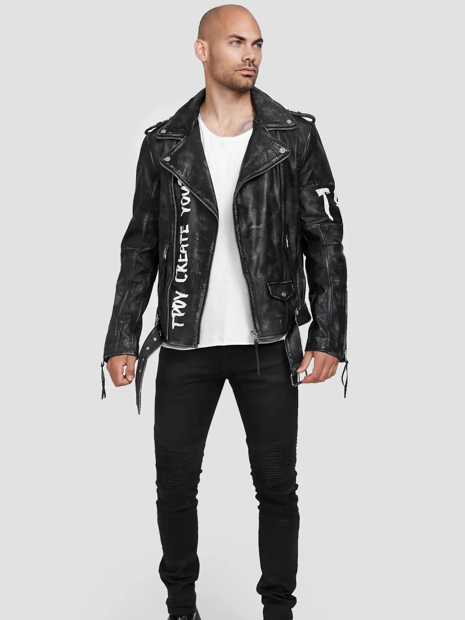 Ein Mann steht vor einem hellgrauen Hintergrund und trägt die Russell Lederjacke mit Print aus Ziegenleder von trueprodigy, gepaart mit einem weißen T-Shirt, schwarzen Jeans und schwarzen Schuhen. Seine Hände liegen entspannt an seinen Seiten, während er leicht nach links schaut.