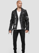 Ein Mann steht vor einem hellgrauen Hintergrund und trägt die Russell Lederjacke mit Print aus Ziegenleder von trueprodigy, gepaart mit einem weißen T-Shirt, schwarzen Jeans und schwarzen Schuhen. Seine Hände liegen entspannt an seinen Seiten, während er leicht nach links schaut.