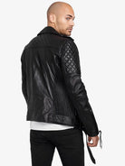 Ein Mann mit rasiertem Kopf modelliert die trueprodigy Simba Bikerlederjacke mit Gürtel in schwarzem Leder mit gesteppten Schultern und Ärmeln. Rückwärts gewandt blickt er über die linke Schulter und trägt eine schwarze Hose und ein weißes Hemd.
