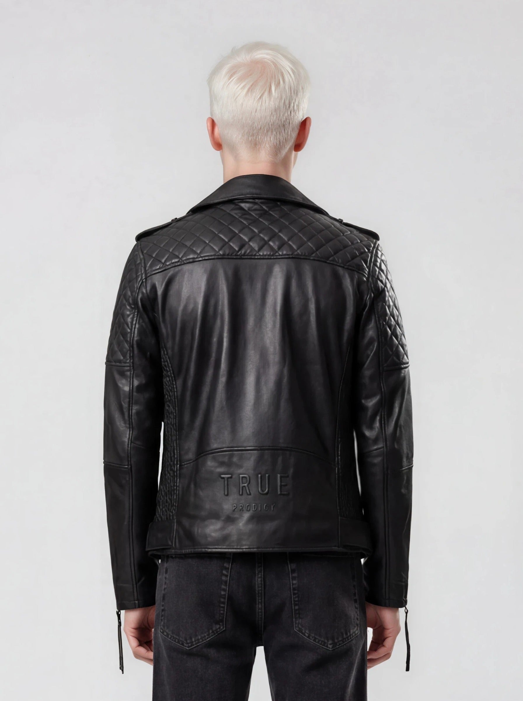 Eine Person mit kurzen, platinblonden Haaren ist von hinten zu sehen. Sie trägt die trueprodigy Biker Lederjacke Herren "Simba" mit Gürtel und schwarze Jeans und steht vor einem schlichten hellgrauen Hintergrund.