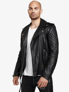Ein Mann mit rasiertem Kopf und kurzem Bart trägt die trueprodigy Simba Bikerlederjacke mit Gürtel in Schwarz über einem weißen T-Shirt und einer schwarzen Hose und steht selbstbewusst vor einem hellen Hintergrund, um diese stilvolle Herrenjacke zu präsentieren.