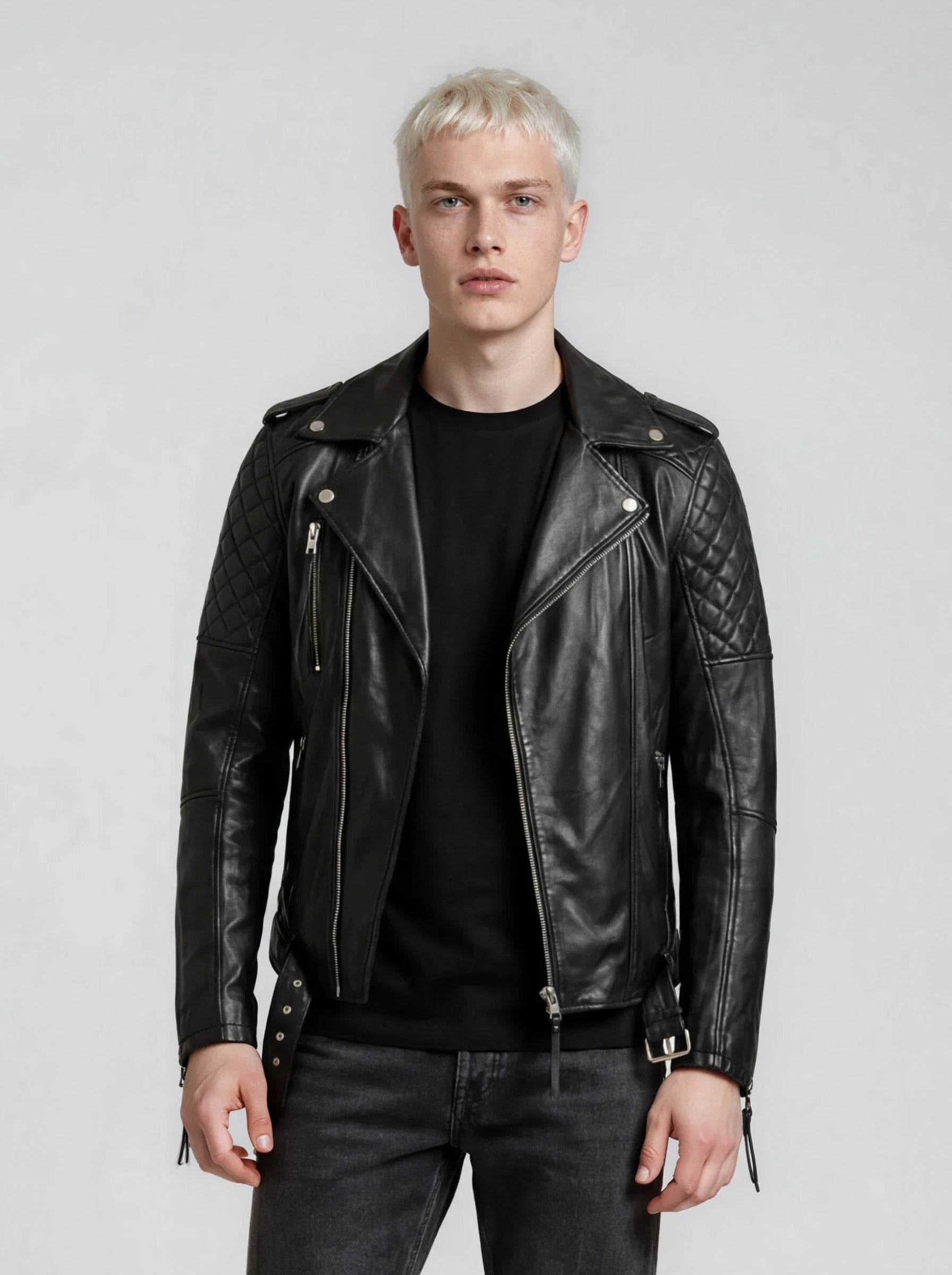 Eine Person mit kurzen blonden Haaren trägt eine trueprodigy Biker Lederjacke Herren "Simba" mit Gürtel über einem schwarzen Hemd und einer dunklen Jeans und steht vor einem schlichten hellgrauen Hintergrund.