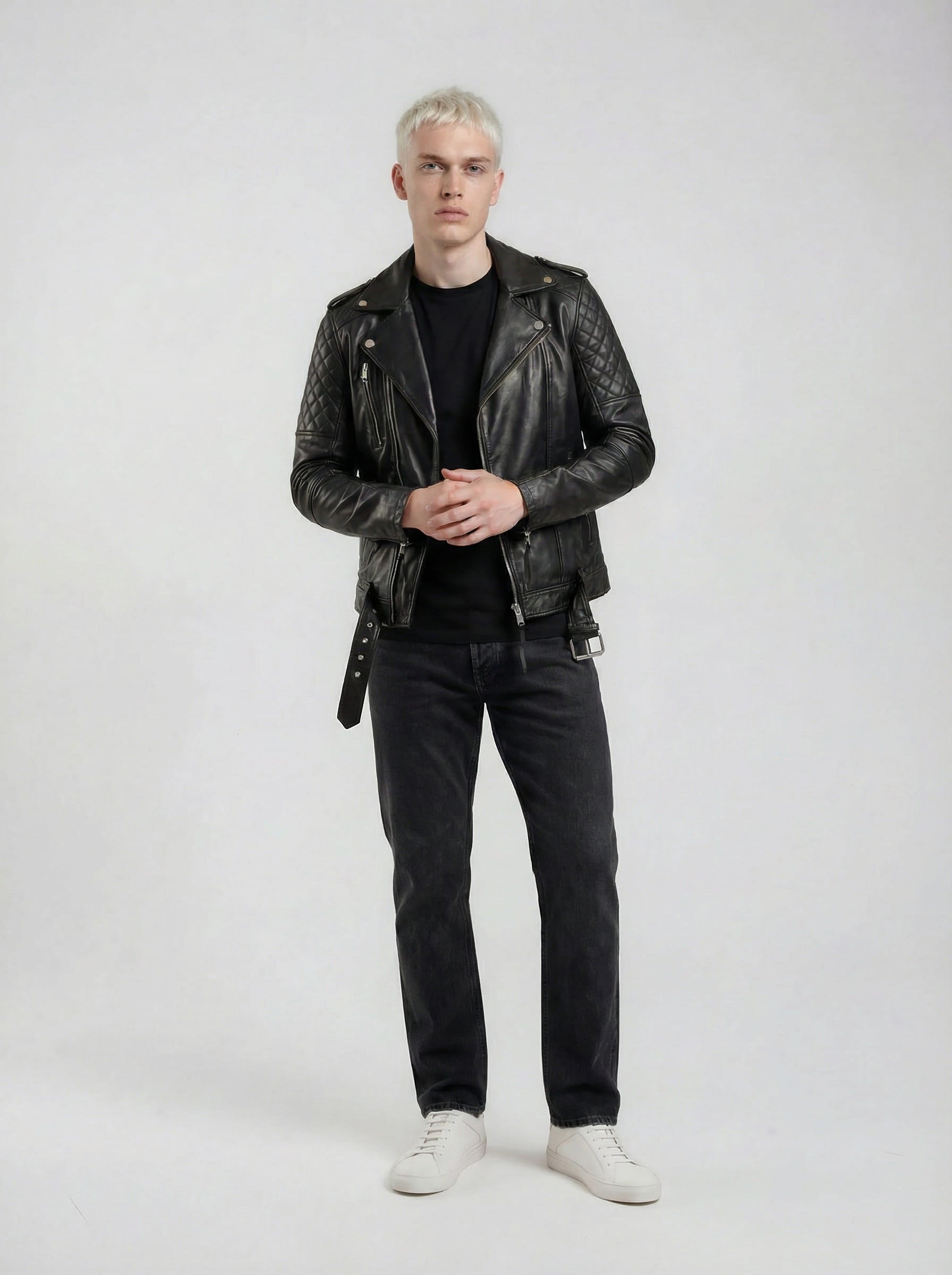 Eine Person mit kurzen, hellen Haaren steht vor einem schlichten Hintergrund und trägt eine trueprodigy Biker Lederjacke Herren "Simba" mit Gürtel, ein schwarzes Hemd, dunkle Jeans und weiße Turnschuhe. Ihre Hände sind auf Hüfthöhe ineinander verschränkt.