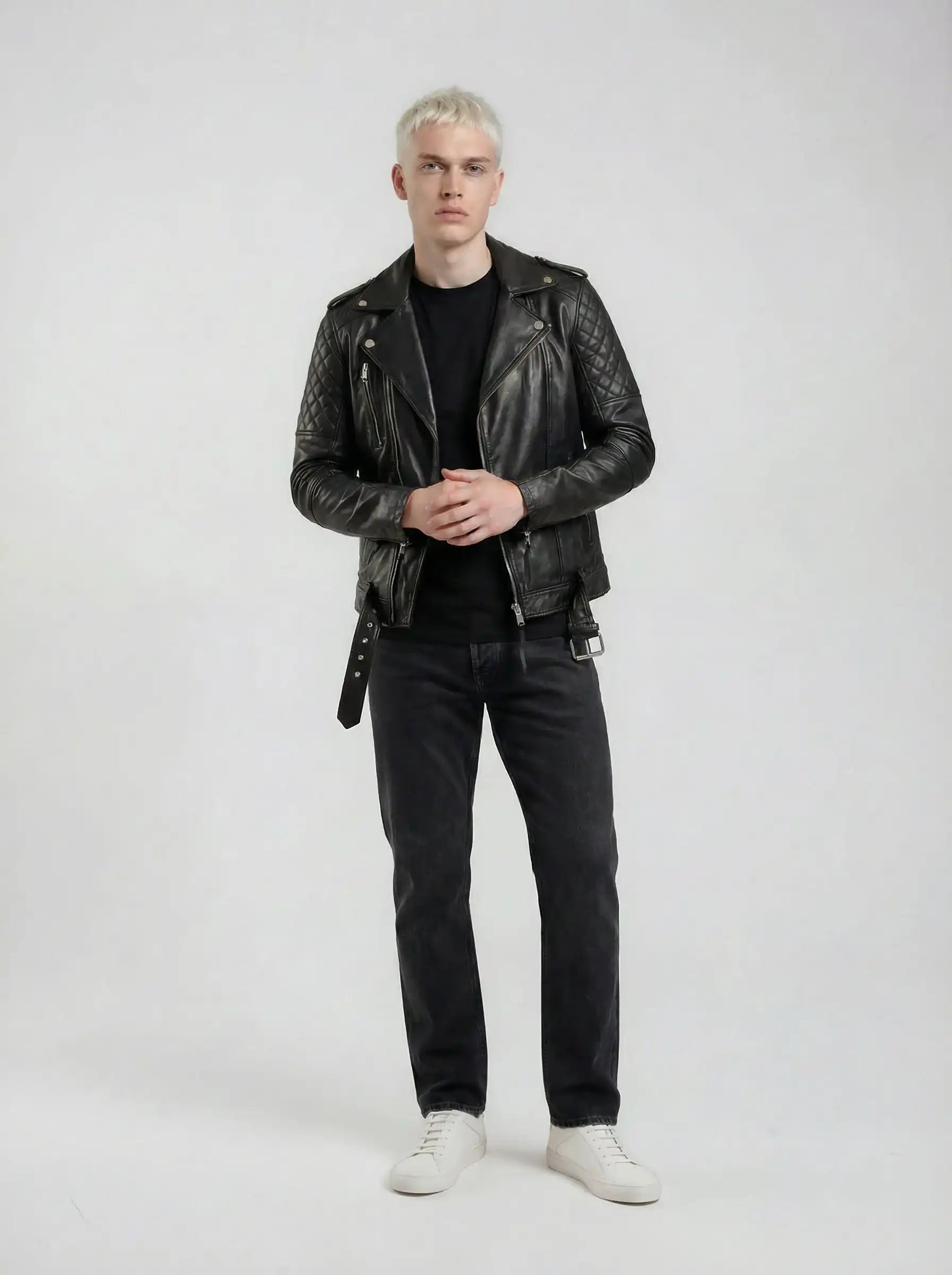 Eine Person mit kurzen, hellen Haaren steht vor einem schlichten Hintergrund und trägt eine trueprodigy Biker Lederjacke Herren "Simba" mit Gürtel, ein schwarzes Hemd, dunkle Jeans und weiße Turnschuhe. Ihre Hände sind auf Hüfthöhe ineinander verschränkt.