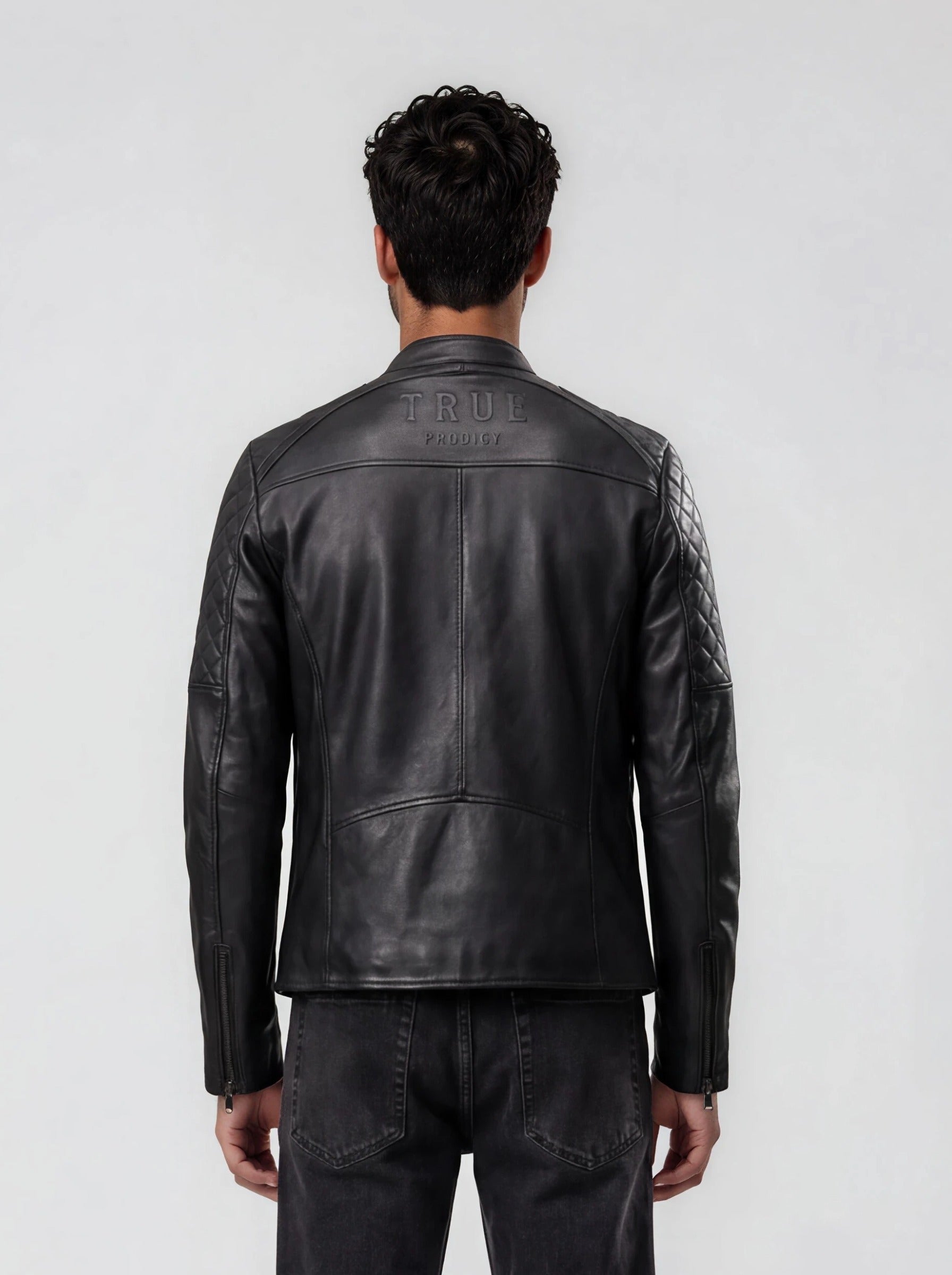 Ein Mann blickt in einer schwarzen trueprodigy Racing Lederjacke Herren "Franky" mit Rautensteppung und dunkler Jeans von der Kamera weg. Auf dem oberen Rücken befindet sich ein auffälliges Branding. Der Hintergrund ist einfarbig, hellgrau.