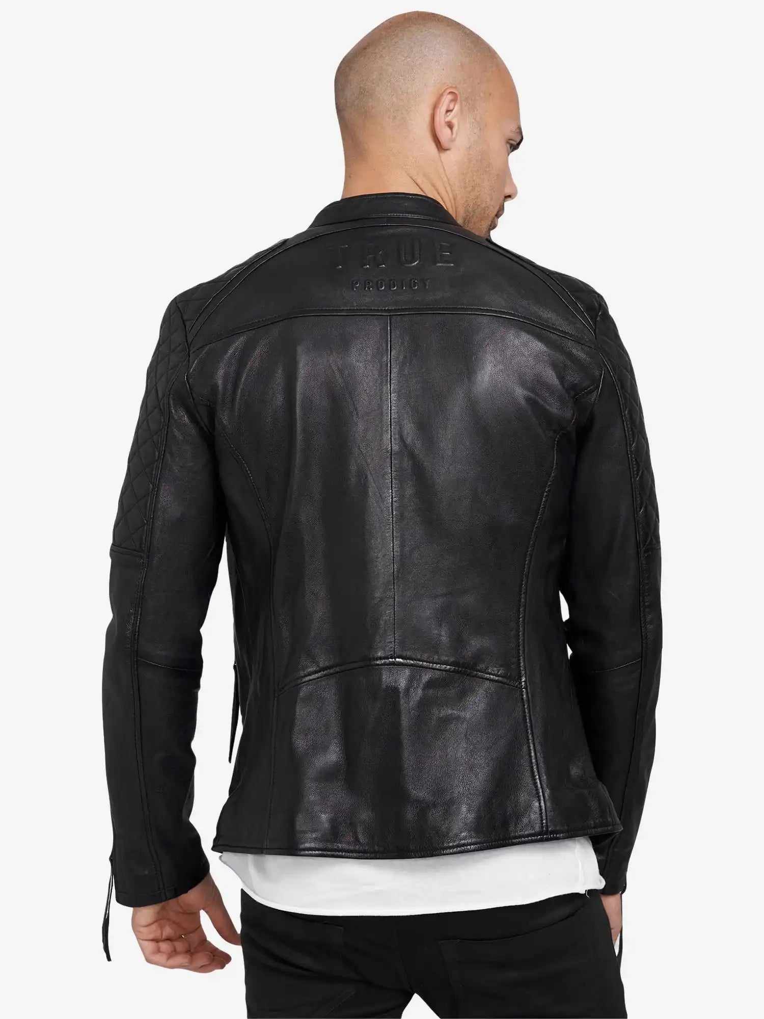 Von hinten ist eine Person mit rasiertem Kopf zu sehen, die eine schwarze Franky Lederjacke mit Stehkragen von trueprodigy mit gesteppten Ärmeln und Schultern, dem eingeprägten Markennamen auf dem oberen Rücken, und einem weißen Hemd darunter trägt.