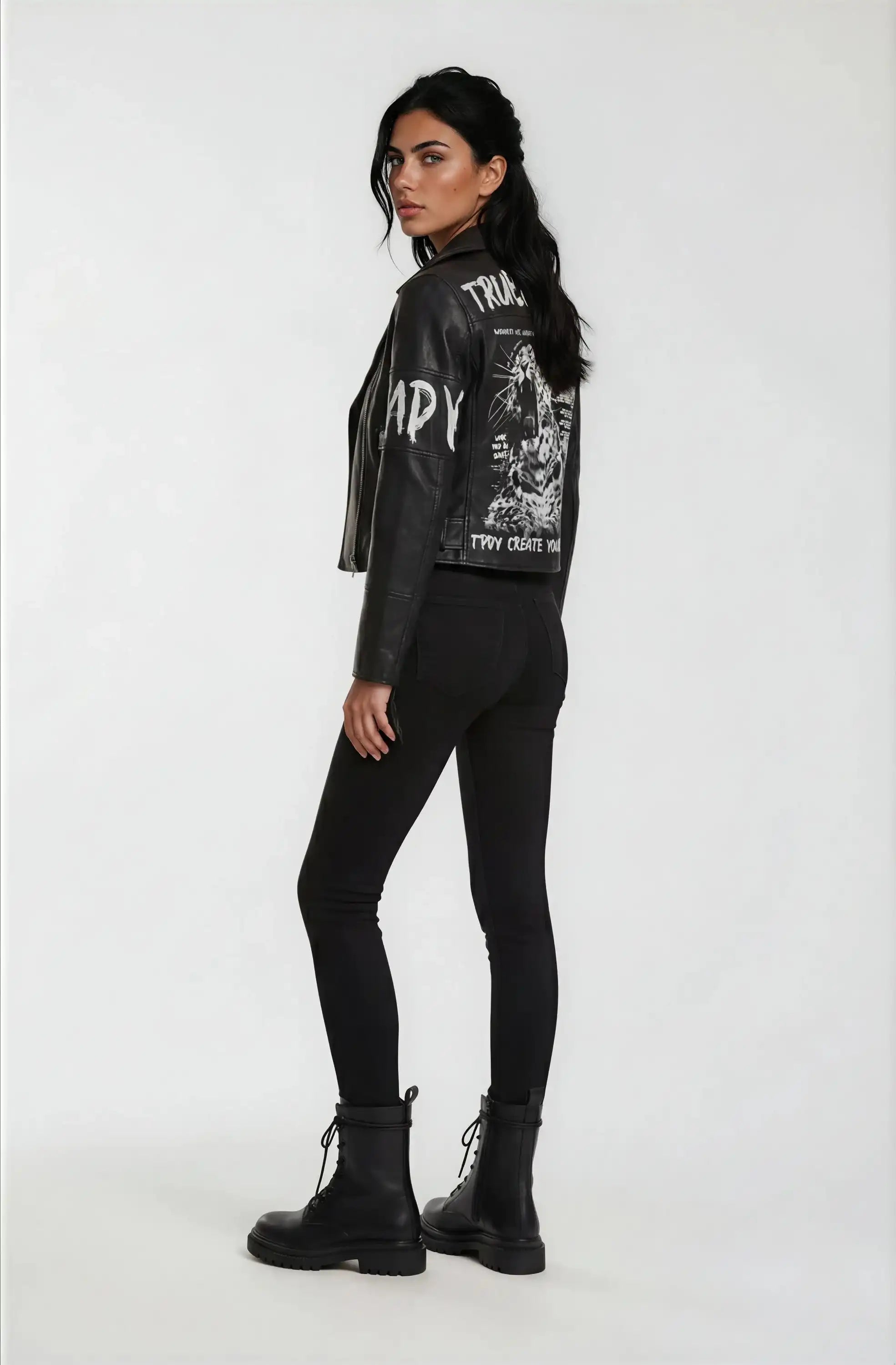 Eine Frau steht in Dreiviertel-Pose mit Blick nach links und trägt die trueprodigy Lederjacke mit Druck mit weißer Grafik auf dem Rücken, gepaart mit schwarzen Skinny Jeans und Schnürstiefeln vor einem schlichten weißen Hintergrund.