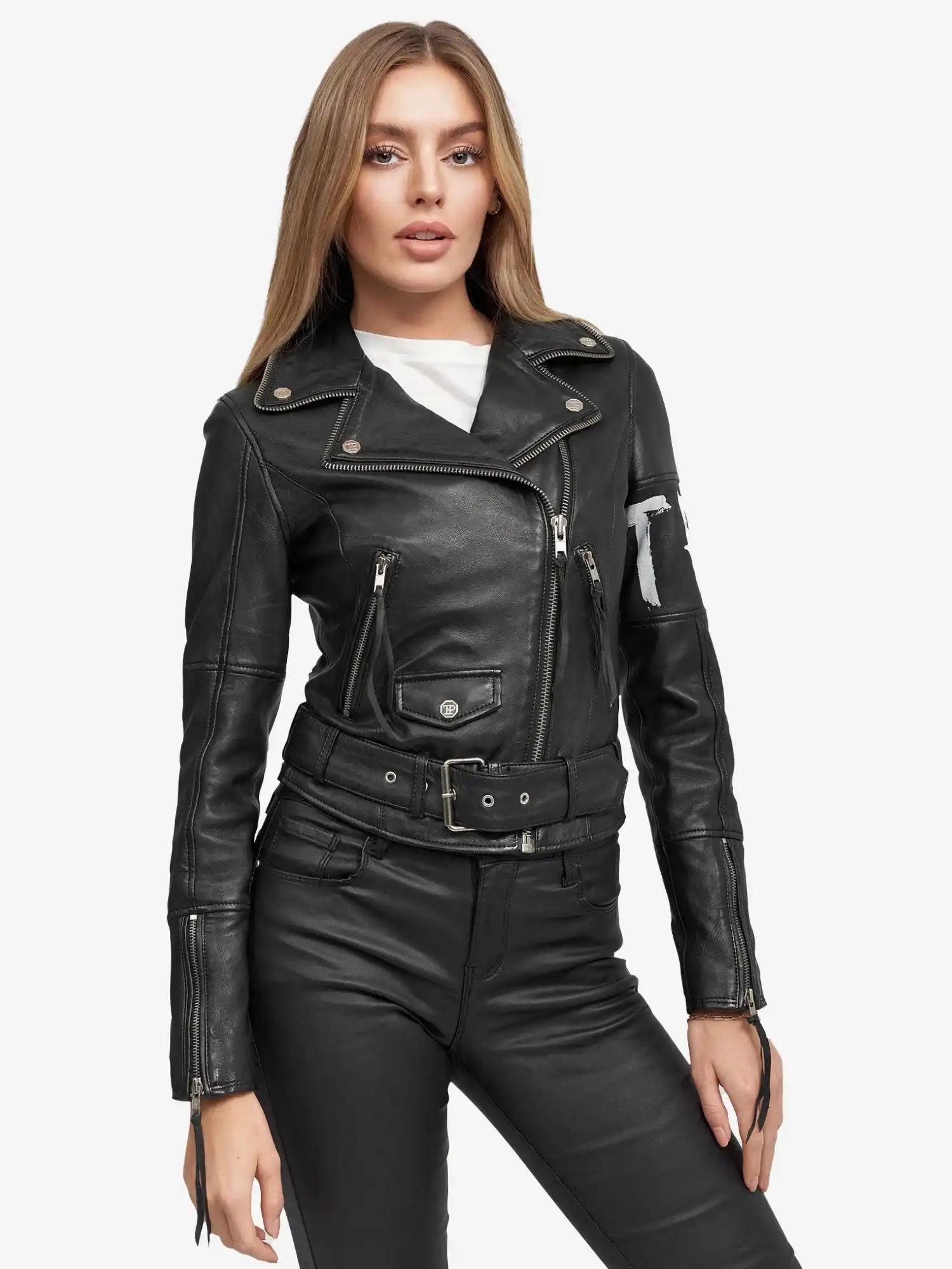 Eine Frau mit langem, glattem hellbraunem Haar trägt die Laila Lederjacke mit Druck von trueprodigy - eine schwarze Biker-Lederjacke über einem weißen Oberteil und einer schwarzen Lederhose - und steht mit dem Gesicht nach vorne vor einem schlichten weißen Hintergrund.