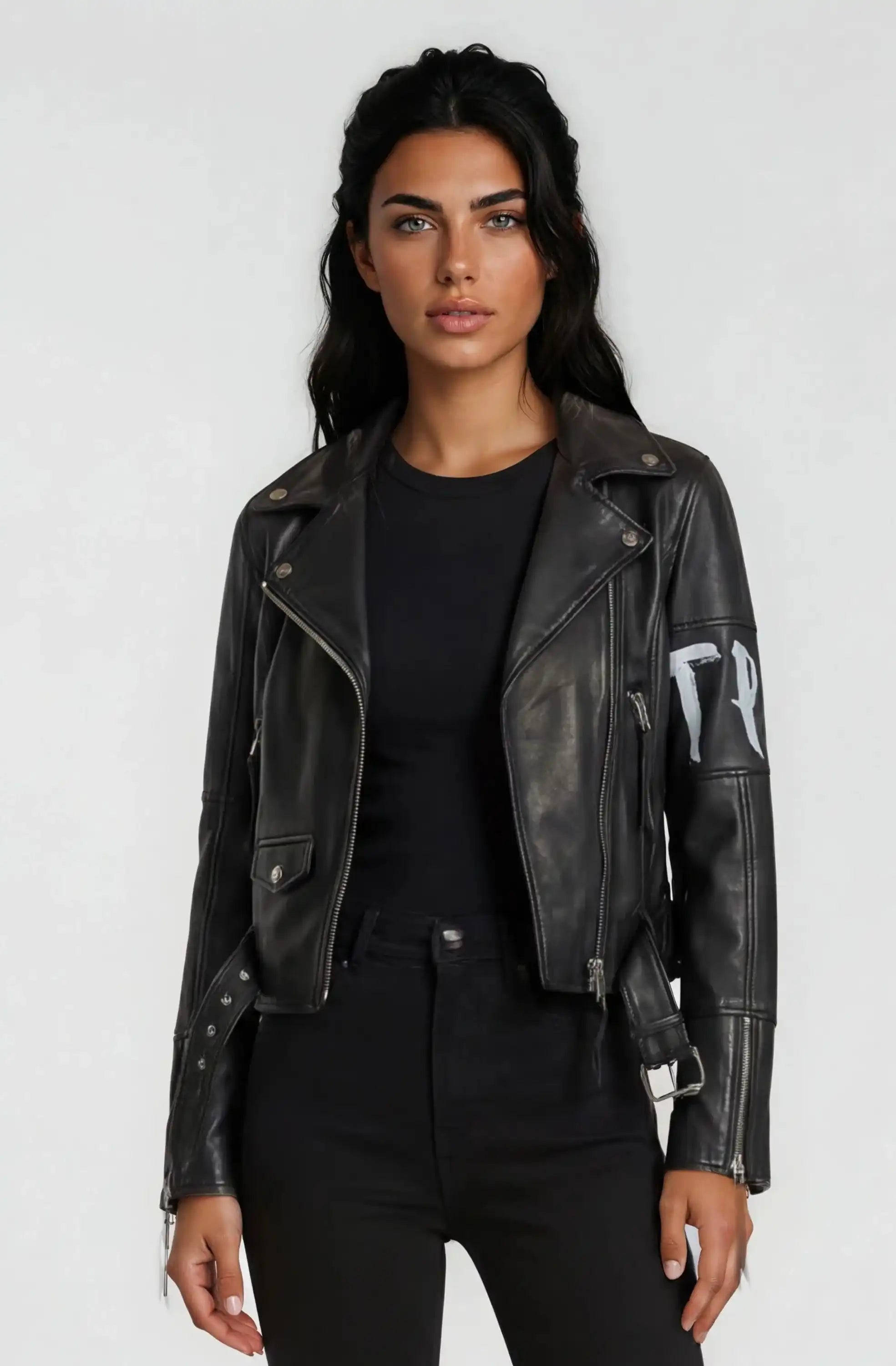 Eine Person mit langen dunklen Haaren trägt die trueprodigy Lederjacke mit Druck über einem schwarzen Hemd und einer schwarzen Hose, die vor einem schlichten hellgrauen Hintergrund steht.