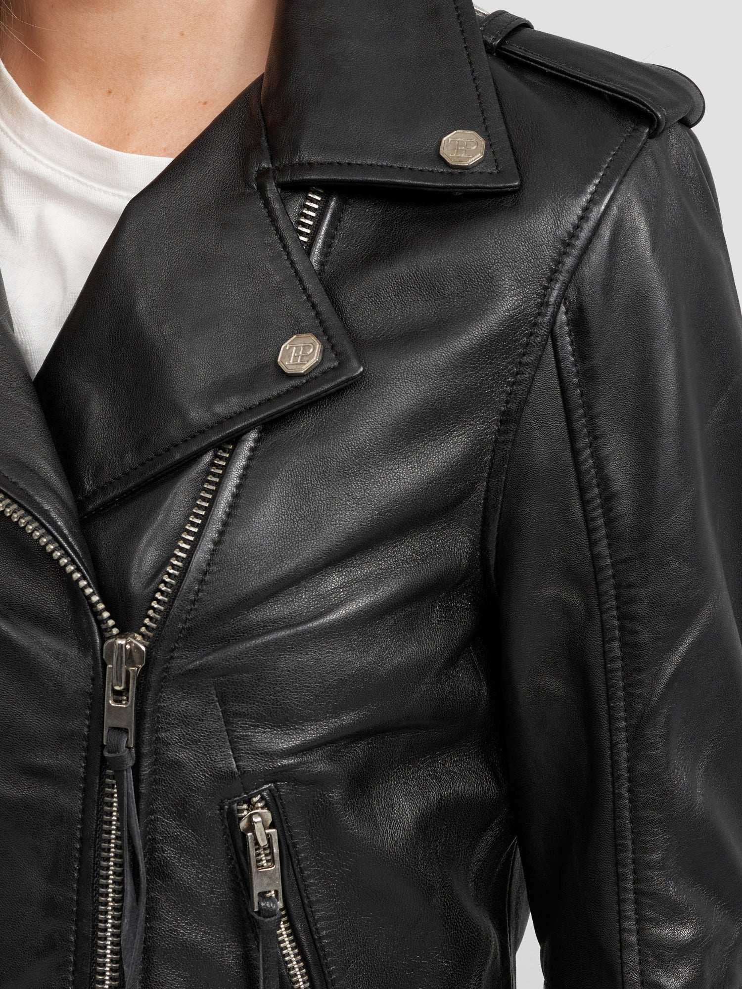 Nahaufnahme der Milena Lederjacke mit Gürtel von trueprodigy, einer schwarzen Damen-Lederjacke mit silbernen Reißverschlüssen und Druckknöpfen, gestylt über einem weißen T-Shirt. Die Aufnahme konzentriert sich auf den oberen Brust- und Schulterbereich.