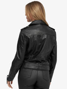 Eine Person mit langen, hellbraunen Haaren wird von hinten gezeigt, die die Trueprodigy Milena Lederjacke mit Gürtel und eine schwarze Hose trägt. Der Hintergrund ist einfarbig und hell.