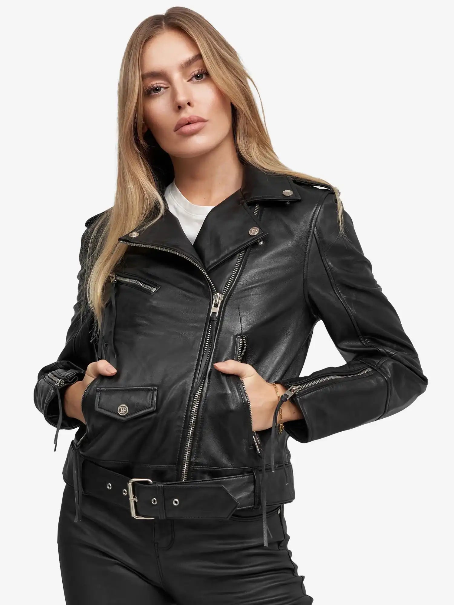 Eine Person mit langen blonden Haaren trägt die Milena Lederjacke mit Gürtel von trueprodigy über einem weißen Hemd. Die schwarze Jacke hat silberne Reißverschlüsse, einen Gürtel und einen Druckknopfkragen. Sie stehen vor einem schlichten hellen Hintergrund, die Hände in den Taschen.