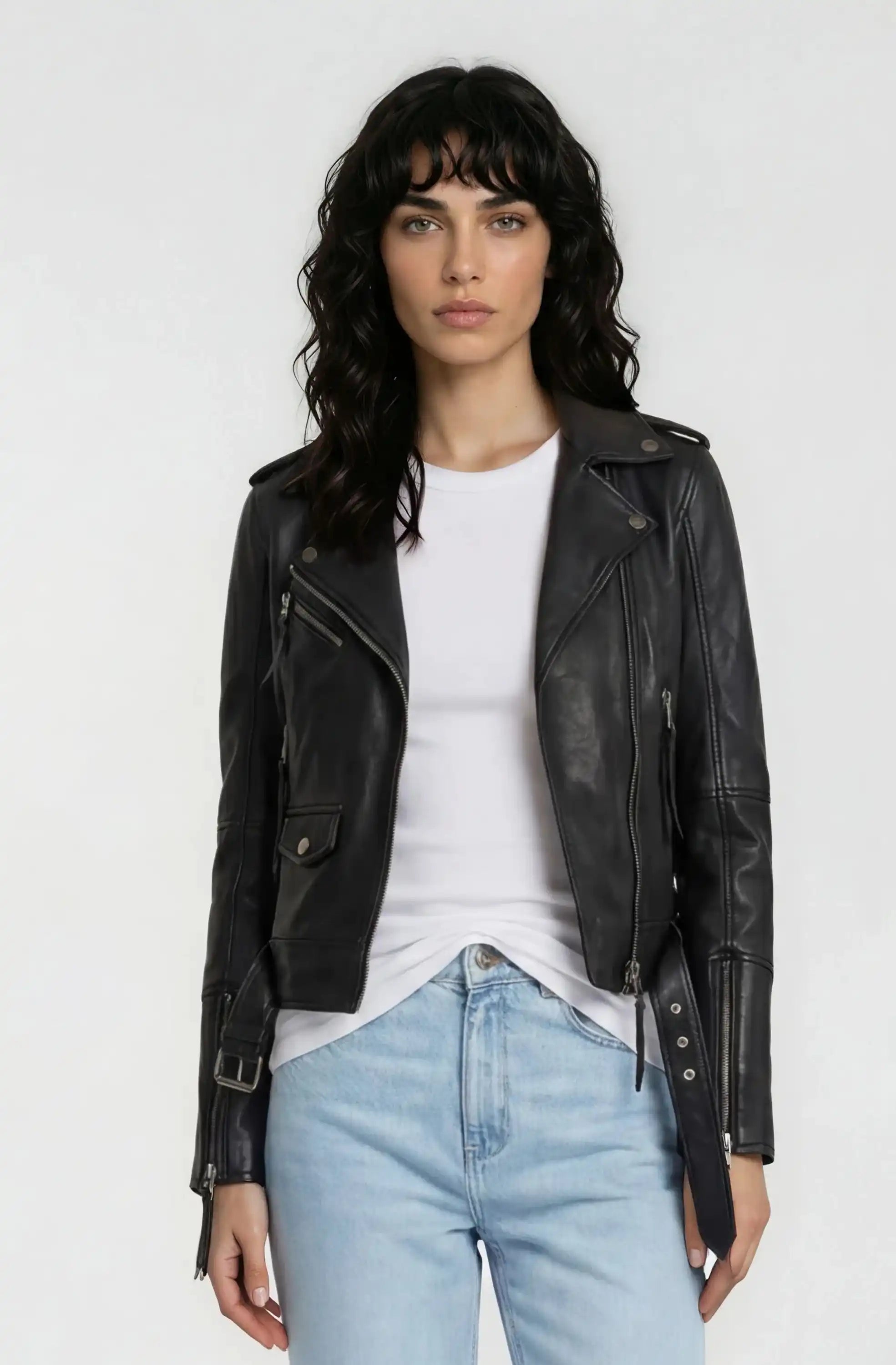 Eine Frau mit gewelltem dunklem Haar trägt die trueprodigy Lederjacke mit Gürtel über einem weißen T-Shirt und hellblauen Jeans und steht mit dem Gesicht nach vorne vor einem schlichten weißen Hintergrund.