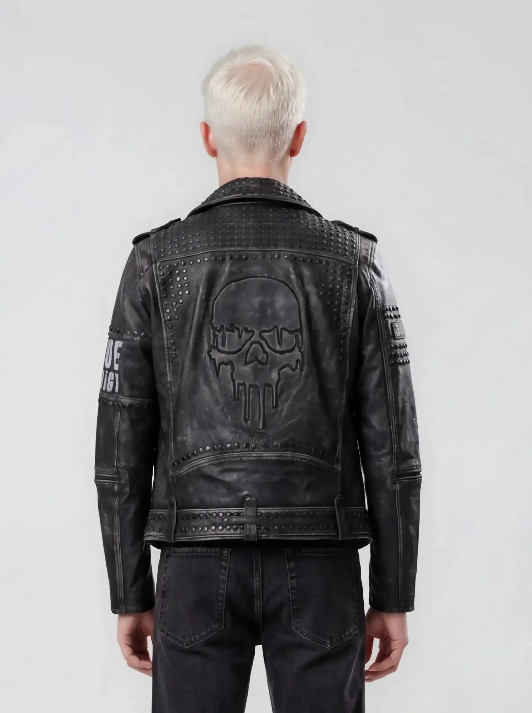 Eine Person mit kurzem, platinblondem Haar steht mit dem Rücken zur Kamera und trägt die trueprodigy Biker Lederjacke Herren "Dwayne" mit Nieten & Print im coolen Biker-Look und kombiniert mit schwarzen Jeans.