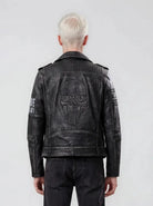 Eine Person mit kurzem, platinblondem Haar steht mit dem Rücken zur Kamera und trägt die trueprodigy Biker Lederjacke Herren "Dwayne" mit Nieten & Print im coolen Biker-Look und kombiniert mit schwarzen Jeans.