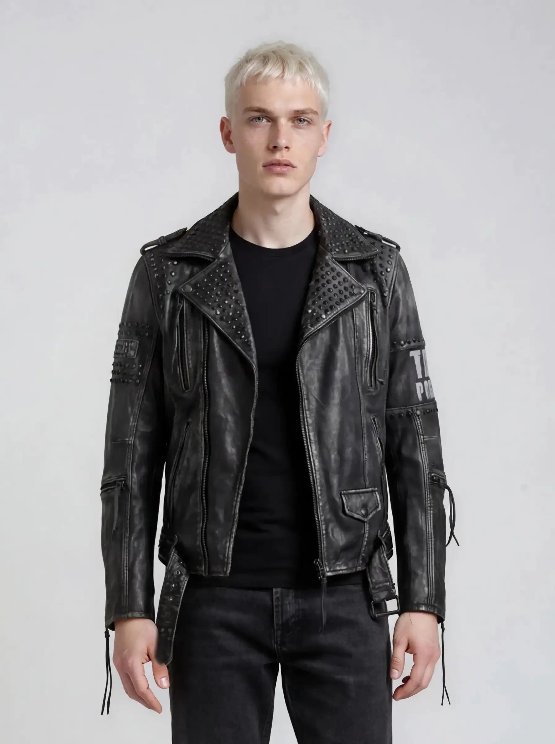Ein junger Mann mit kurzen blonden Haaren trägt die trueprodigy Biker Lederjacke Herren "Dwayne" mit Nieten & Print über schwarzem T-Shirt und Jeans, steht vor hellgrauem Hintergrund und blickt neutral nach vorne.