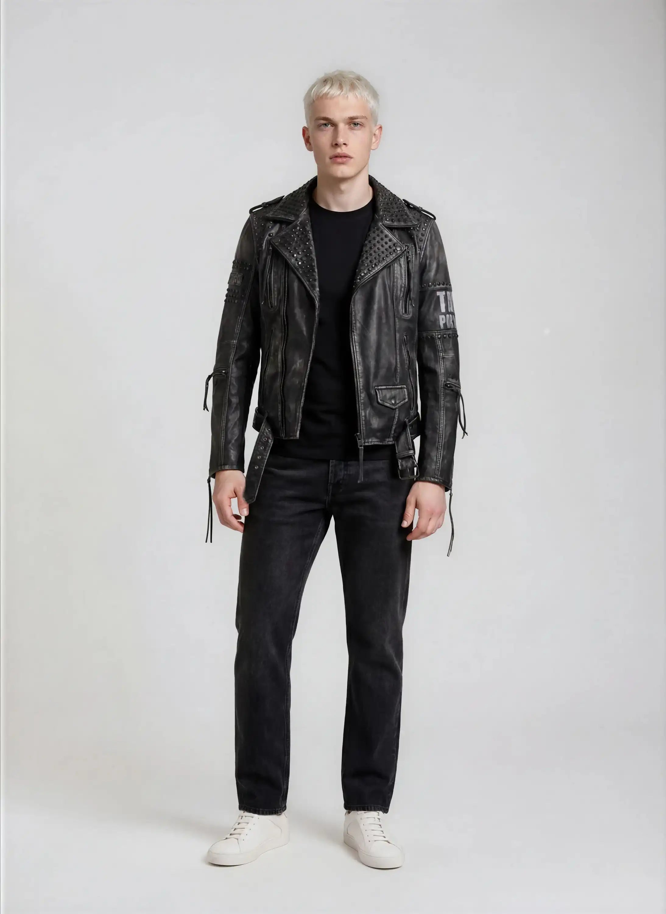 Eine Person mit kurzen blonden Haaren steht vor einem schlichten Hintergrund, trägt die trueprodigy Biker Lederjacke Herren "Dwayne" mit Nieten & Print, ein schwarzes T-Shirt, dunkle Jeans und weiße Turnschuhe und blickt mit neutralem Blick nach vorne.
