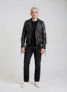 Eine Person mit kurzen blonden Haaren steht vor einem schlichten Hintergrund, trägt die trueprodigy Biker Lederjacke Herren "Dwayne" mit Nieten & Print, ein schwarzes T-Shirt, dunkle Jeans und weiße Turnschuhe und blickt mit neutralem Blick nach vorne.