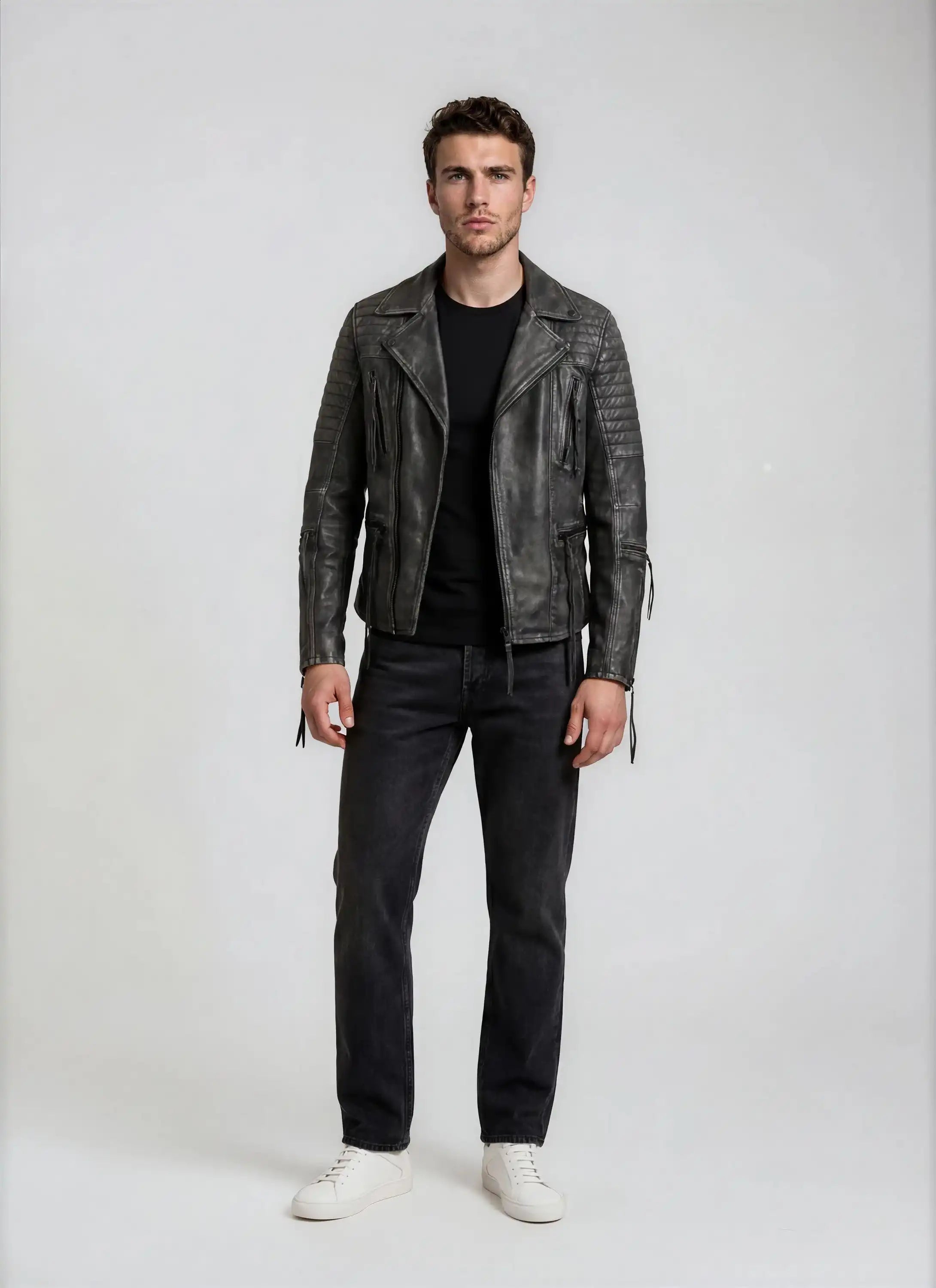 Ein Mann steht mit dem Gesicht zur Kamera vor einem schlichten Hintergrund und trägt die trueprodigy Vintage Biker Lederjacke Herren "Bryan" mit Steppung, ein schwarzes T-Shirt, dunkle Jeans und weiße Turnschuhe. Seine Arme liegen entspannt an den Seiten und sein Gesichtsausdruck ist neutral.