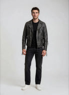 Ein Mann steht mit dem Gesicht zur Kamera vor einem schlichten Hintergrund und trägt die trueprodigy Vintage Biker Lederjacke Herren "Bryan" mit Steppung, ein schwarzes T-Shirt, dunkle Jeans und weiße Turnschuhe. Seine Arme liegen entspannt an den Seiten und sein Gesichtsausdruck ist neutral.