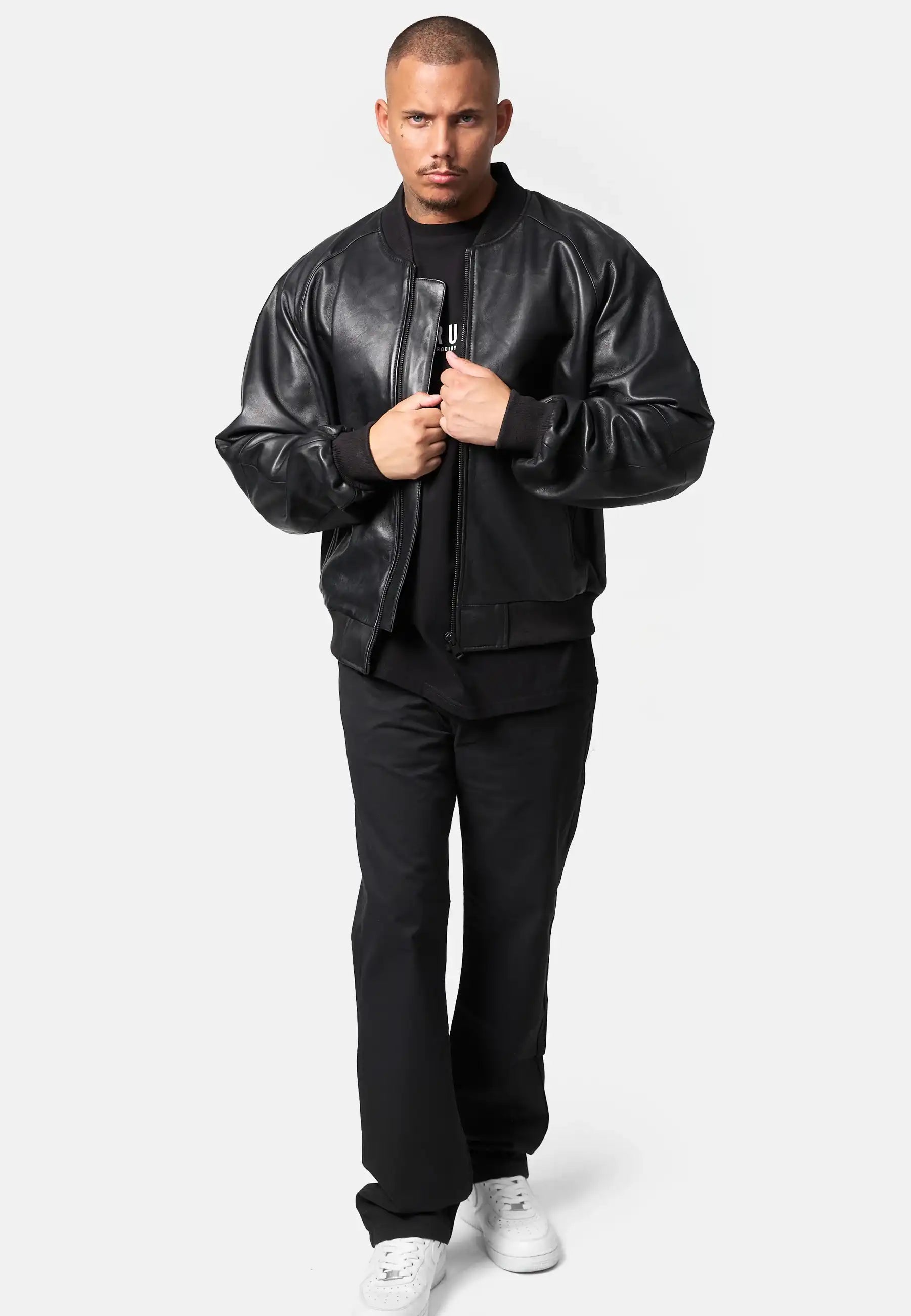 Ein Mann mit kurzen Haaren trägt eine trueprodigy Filip Oversized Bomber-Lederjacke über einem schwarzen Hemd und einer Hose, gepaart mit weißen Sneakers. Er steht vor einem schlichten weißen Hintergrund und hält die Vorderseite seiner Jacke mit beiden Händen fest.