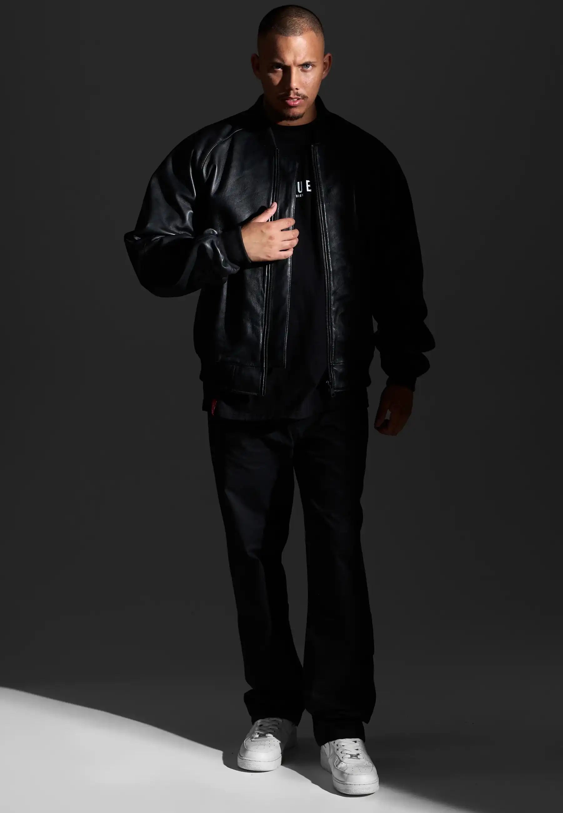Ein Mann steht vor einem dunklen Hintergrund und trägt die trueprodigy Filip Oversized Bomber-Lederjacke, eine schwarze Hose und ein schwarzes Hemd mit weißem Schriftzug, gepaart mit weißen Sneakers. Er blickt nach vorne und hält eine Hand an den Reißverschluss seiner Jacke.