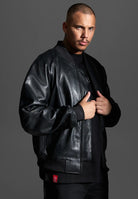 Ein Mann mit kurzen Haaren und gestutztem Schnurrbart trägt die trueprodigy Filip Oversized Bomber-Lederjacke über einem schwarzen Hemd, steht vor einem grauen Hintergrund und schaut mit neutralem Blick direkt in die Kamera.