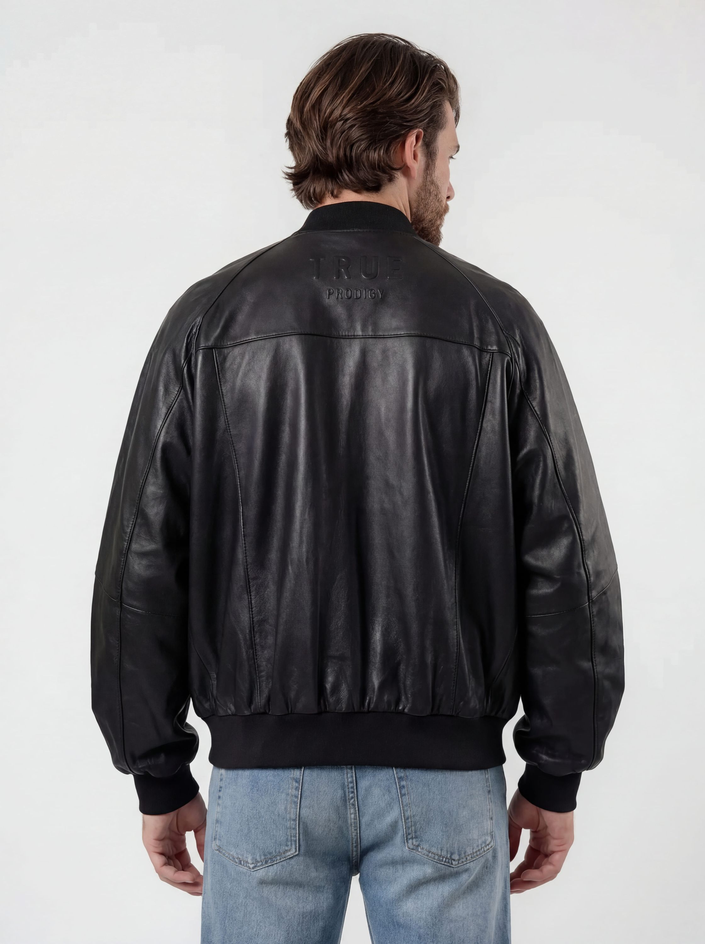 Eine Person mit hellbraunen Haaren trägt die trueprodigy Bomber Lederjacke Herren "Filip" im Oversize-Look und hellblaue Jeans und steht mit dem Rücken zur Kamera vor einem schlichten, hellen Hintergrund.