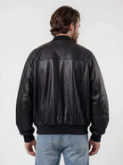 Eine Person mit hellbraunen Haaren trägt die trueprodigy Bomber Lederjacke Herren "Filip" im Oversize-Look und hellblaue Jeans und steht mit dem Rücken zur Kamera vor einem schlichten, hellen Hintergrund.