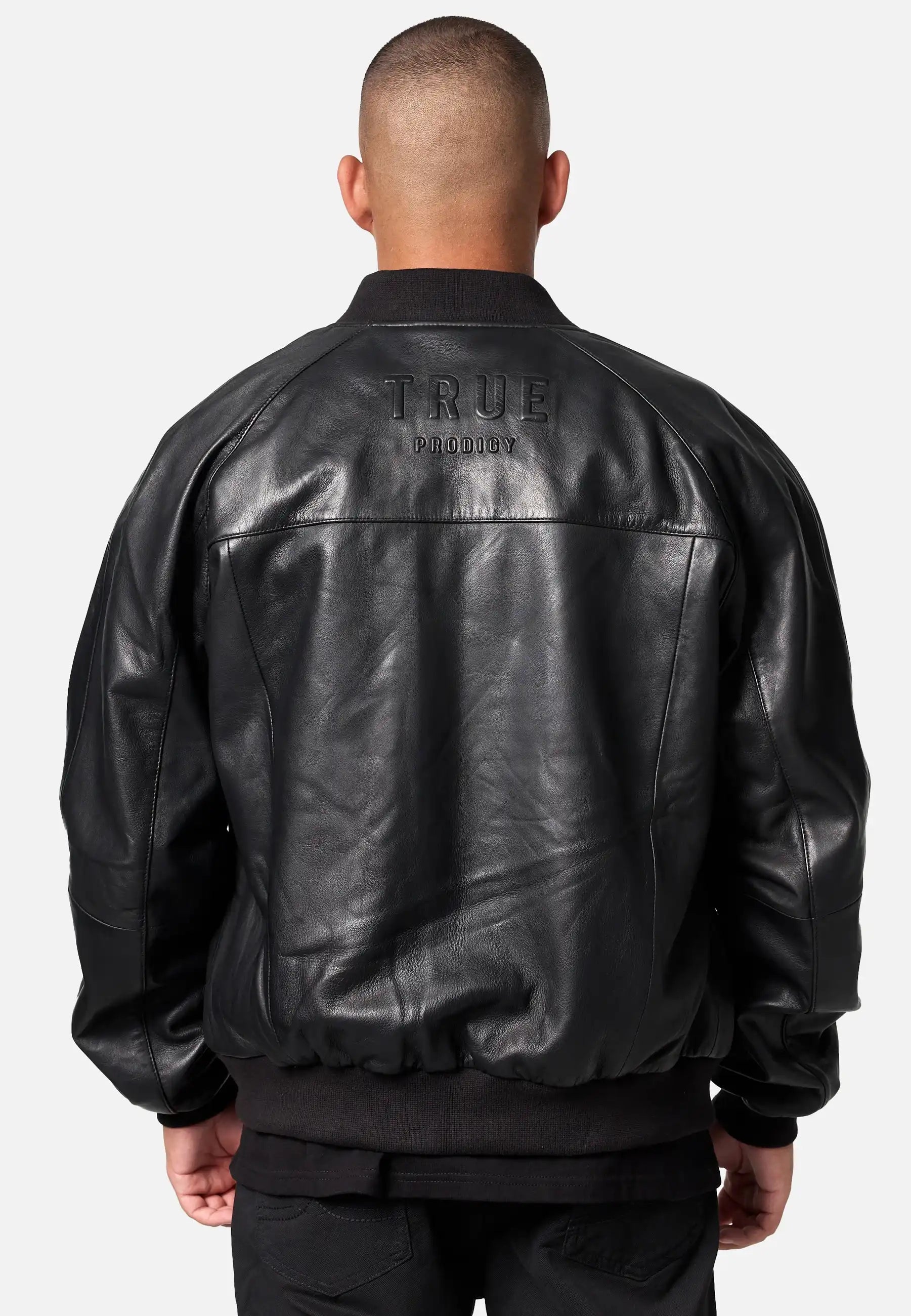 Eine Person steht mit dem Rücken zur Kamera und trägt eine trueprodigy Filip Oversized Bomber-Lederjacke mit dem großen TRUE PRODIGY-Schriftzug auf dem Rücken. Der Hintergrund ist hell und schlicht.